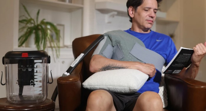 Man seated at home reading with an ICEMAN Clear3 shoulder wrap fitted and the ICEMAN cold therapy unit beside the chair