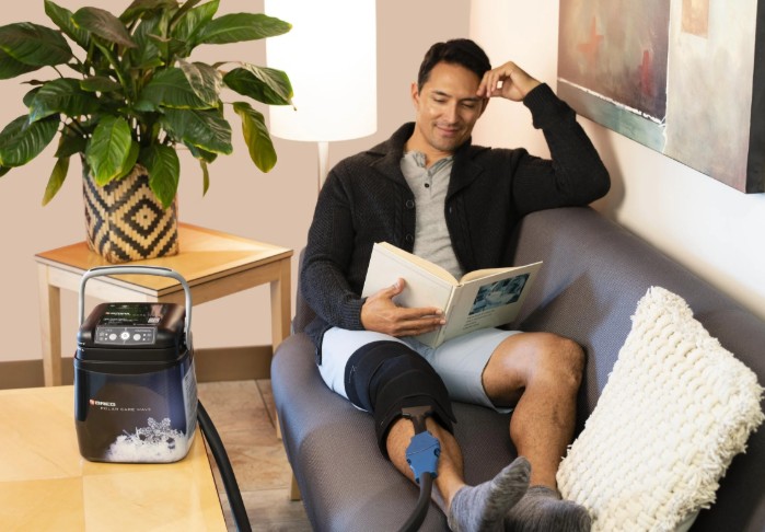 Man relaxing at home reading a book with a Polar Care Wave knee wrap and the Polar Care Wave cold therapy unit on the side table